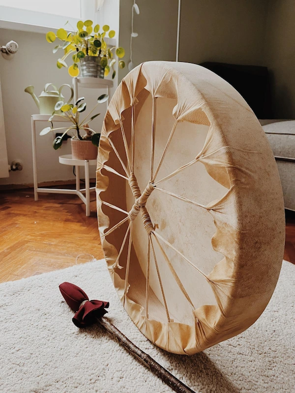 bubanj Šamanski bubanj oslonjen na bijeli tepih u toplom, prirodno uređenom dnevnom boravku, uz drvenu palicu i sobne biljke kraj prozora.