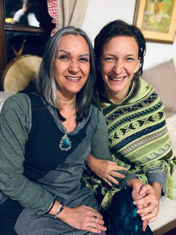 Dvije žene sjede blizu jedna drugoj u zatvorenom prostoru i toplo se smiješe prema kameri. Jedna (Irena) ima dugu sijedu kosu i nosi zeleno-sivu odjeću s ogrlicom s tirkiznim privjeskom, dok druga (Senka) ima smeđu kosu i ogrnuta je zelenim šalom s uzorkom. Drže se za ruke i lagano se naginju jedna prema drugoj, stvarajući ugodnu i nježnu atmosferu.