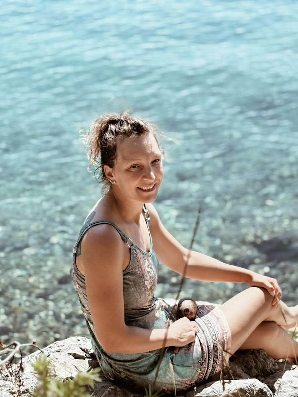 ja-naslovna Žena (Senka) sjedi na stijeni uz kristalno čisto more, nasmiješena i opuštena, okružena prirodom i sunčevim svjetlom.