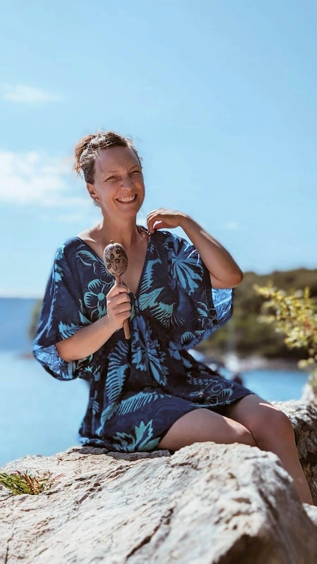 ja_2 Žena (Senka) sjedi na osunčanoj stijeni uz more i vedro se smiješi držeći rezbarenu drvenu šuškalicu. Nosi plavu haljinu s velikim uzorkom tropskog lišća, kosa joj je podignuta, a iza nje se vidi plavo nebo. Prizor odiše radošću i opuštenošću uz vodu.