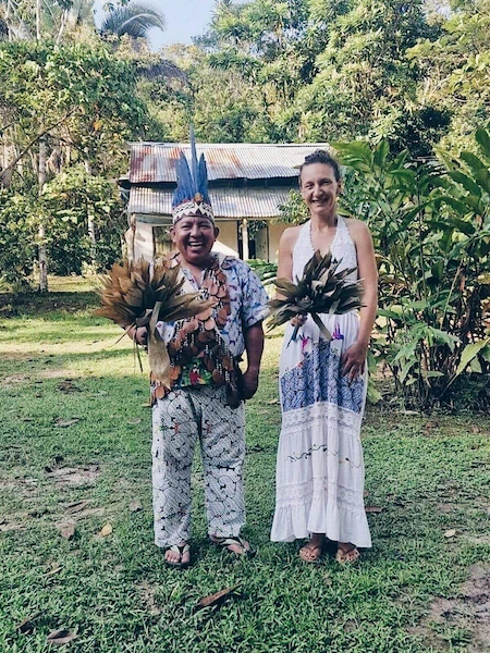 Senka stoji na vani na otvorenom, na travnatoj površini, uz svog učitelja Javiera Arévala Shahuana. Oboje drže snopove lišća koji se koriste u ceremonijalnim praksama. Senka nosi dugu bijelu haljinu s plavim uzorcima, dok šaman nosi šarenu tradicionalnu odjeću s visokim pernatim oglavnikom. Oboje se smiješe, a u pozadini se nalaze bujna zelena tropska vegetacija i mala rustikalna građevina.