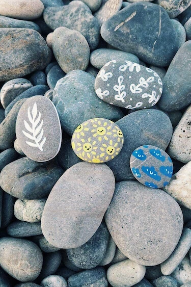 radionica za djecu Glatko morsko kamenje u nijansama sive i plave, na kojem su oslikani jednostavni motivi: bijeli list, sitne grančice, žuti krugovi s nasmiješenim licima i plavi oblaci. Kamenčići su položeni među prirodno kamenje na šljunčanoj podlozi.