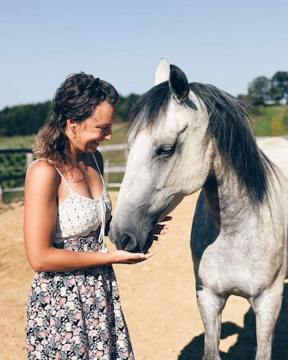 seansa za zivotinje Žena (Senka) nježno drži ruku ispod njuške sivog konja, stojeći nasuprot njega na sunčanom vanjskom prostoru. Oboje djeluju opušteno i povezano, u mirnom prirodnom okruženju s ogradom i zelenilom u pozadini.
